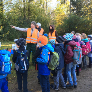 Ein Mann in Warnweste steht vor einer Gruppe von Kindern und deutet in die Ferne.