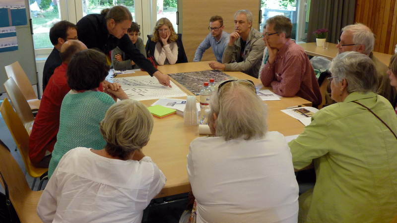 Eine Gruppe von Bürger*innen sitzt um einen großen Tisch auf dem Baupläne der geplanten U1 Oldenfelde liegen. 