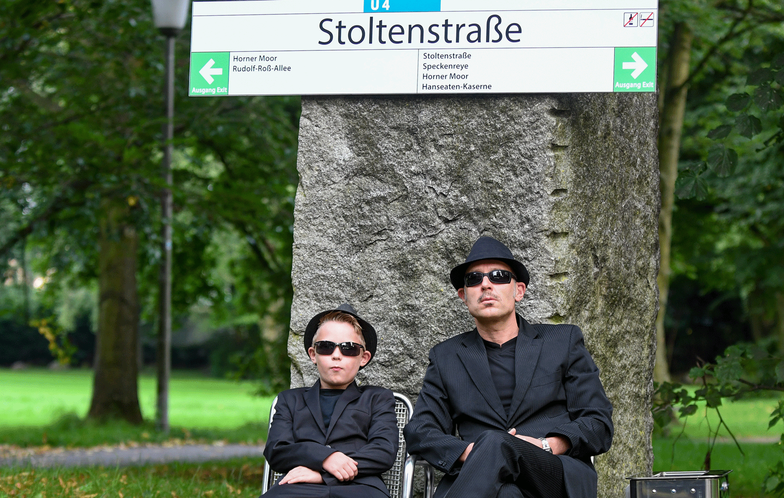 Ein Kind und ein Mann, beide in schwarzen Anzügen und mit Sonnenbrille, sitzen untern einem Haltestellenschild mit der Aufschrift "Stoltenstraße".