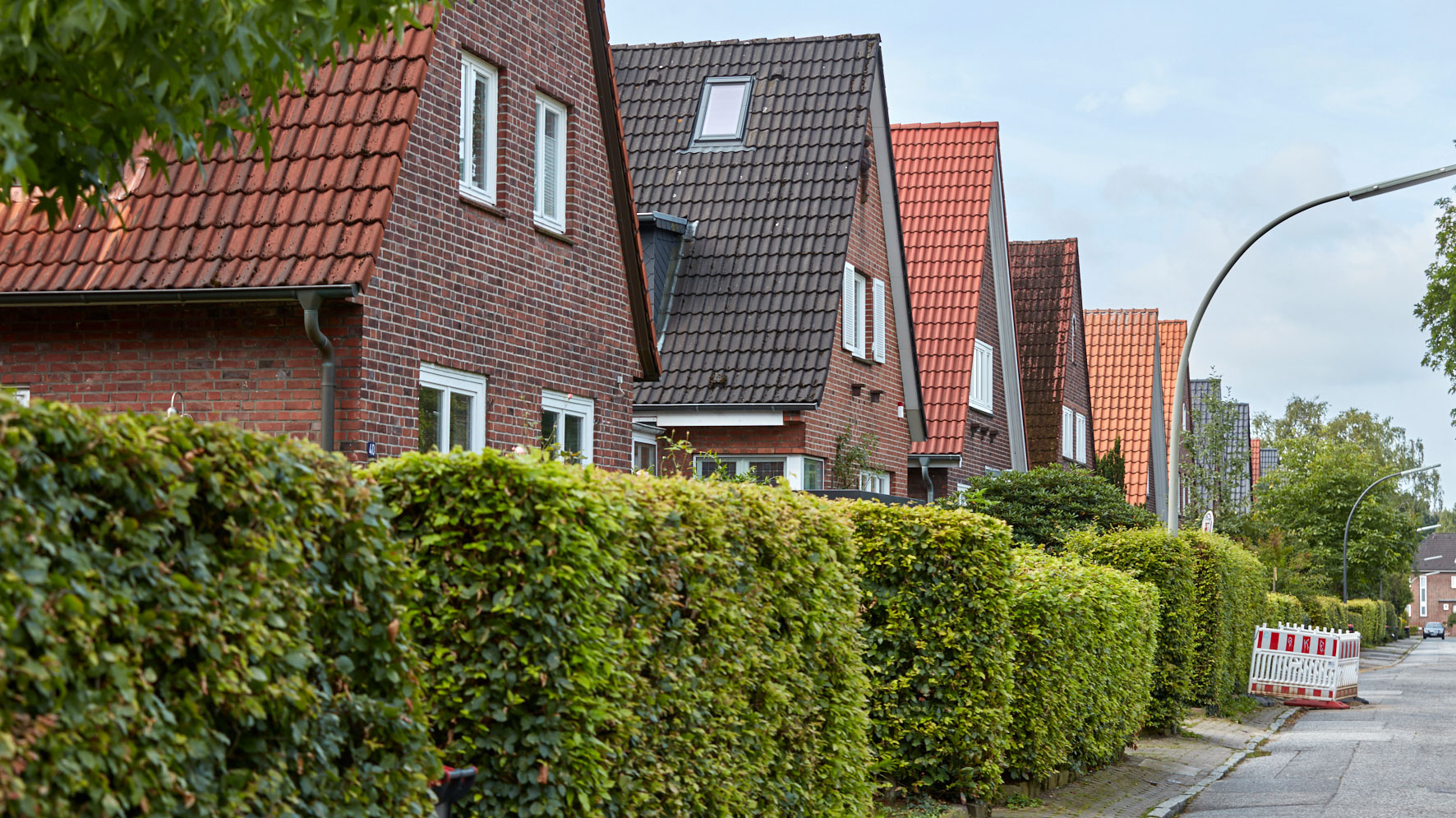 Einfamilienhäuser mit Satteldächern reihen sich hinter grünen Hecken aneinander.