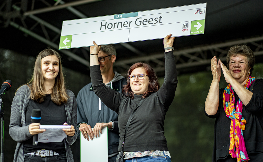 Eine Frau hält ein Haltestellenschild mit dem Namen "Horner Geest" in die Höhe.