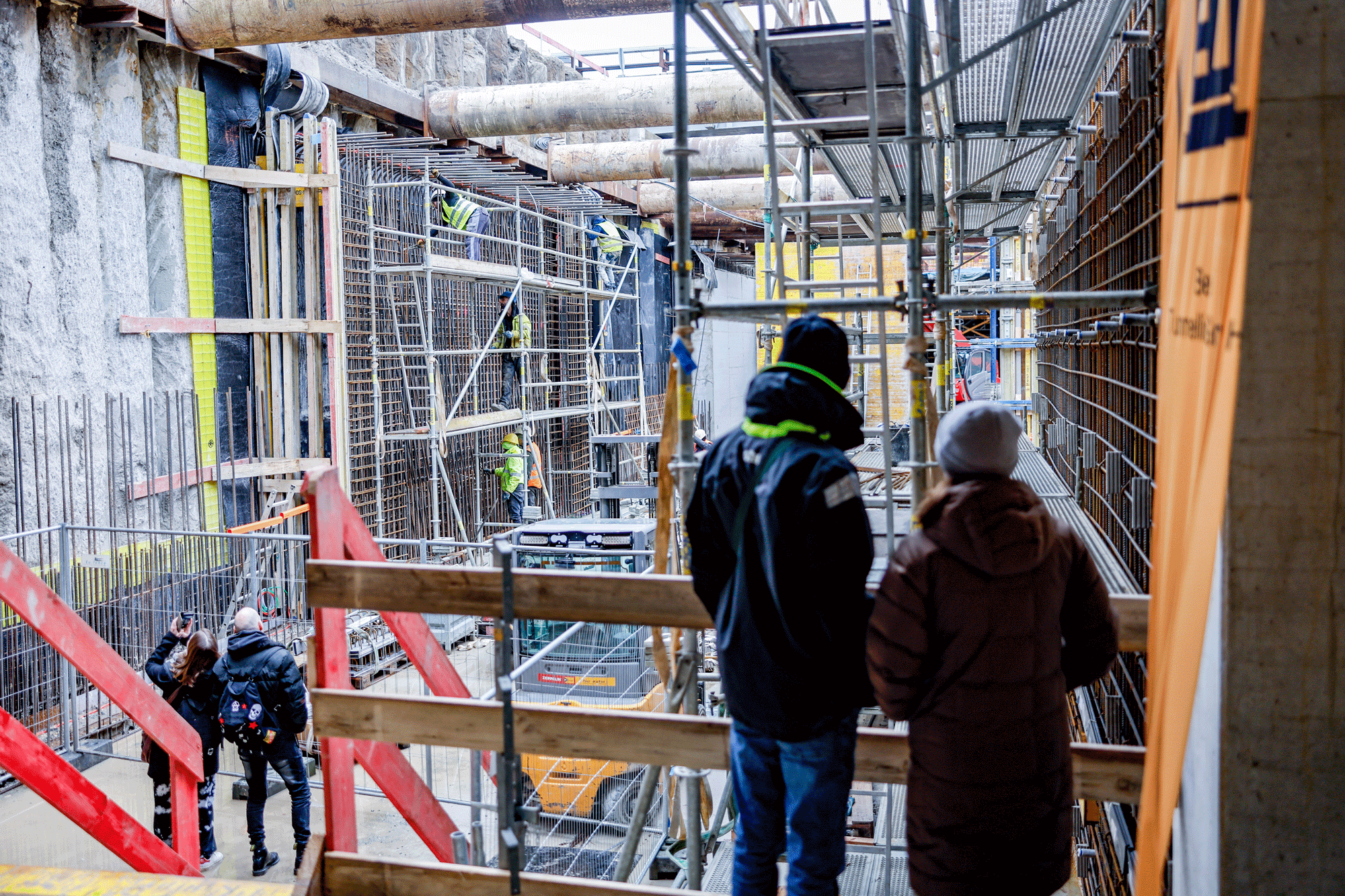 Blick aus der Baugrube in die Baugrube. Zwei Personen schauen von einer Erhöhung in die Baugrube, Im Hintergrund finden Arbeiten statt