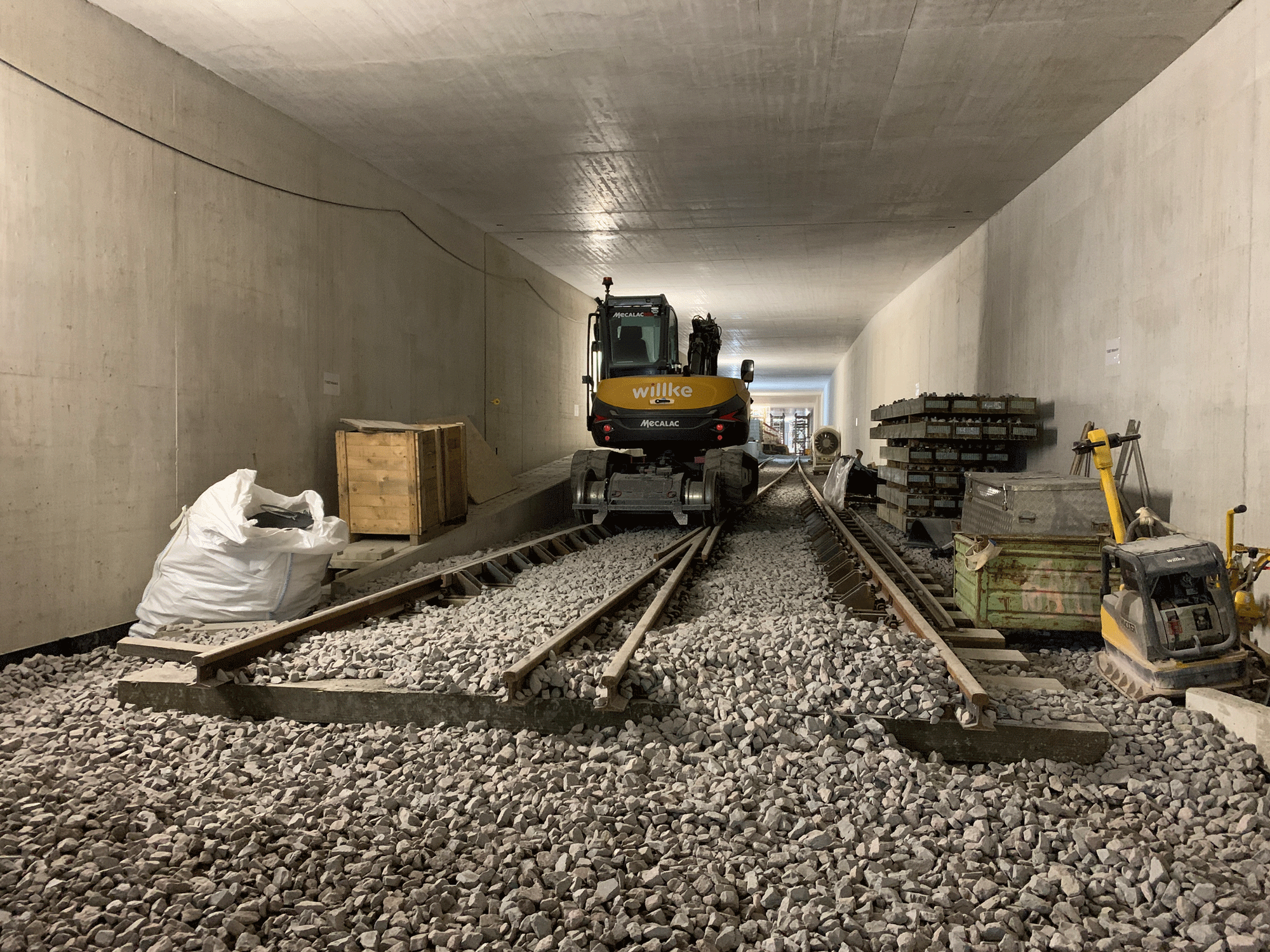 Bild vom fertiggestellten Tunnel Schotter und Teile von Gleisen sind zu sehen, sowie ein Zweiwegebagger auf den Gleisen.