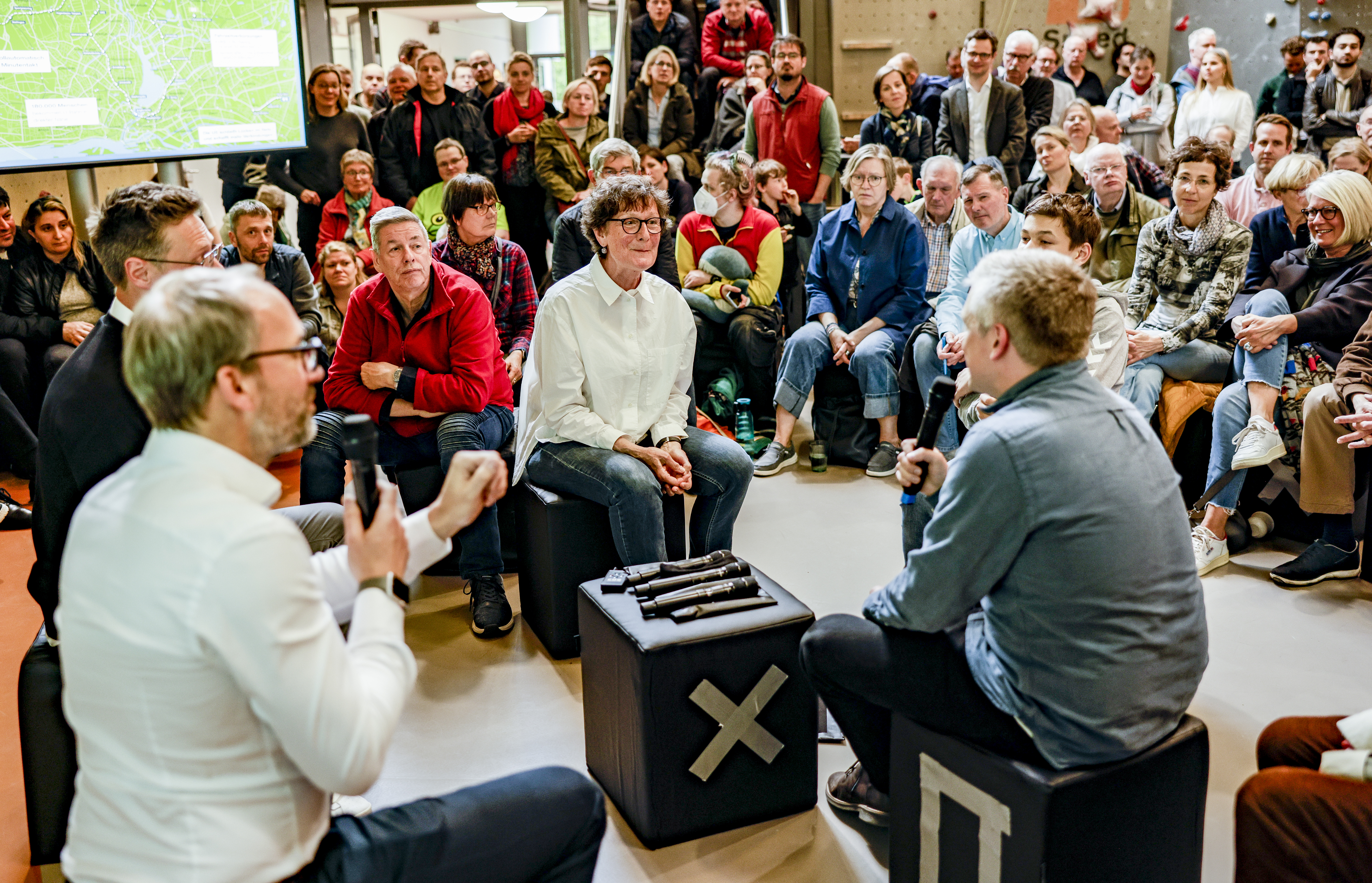 Drei Menschen mit Mikro sitzen in der Mitte, im Hintergrund Publikum und eine Präsentation.