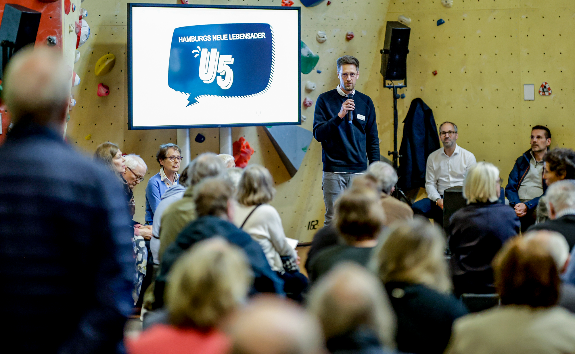 Publikum bei einer Veranstaltung von hinten. Im Fokus ein U5-Sprecher neben einer Präsentation. 