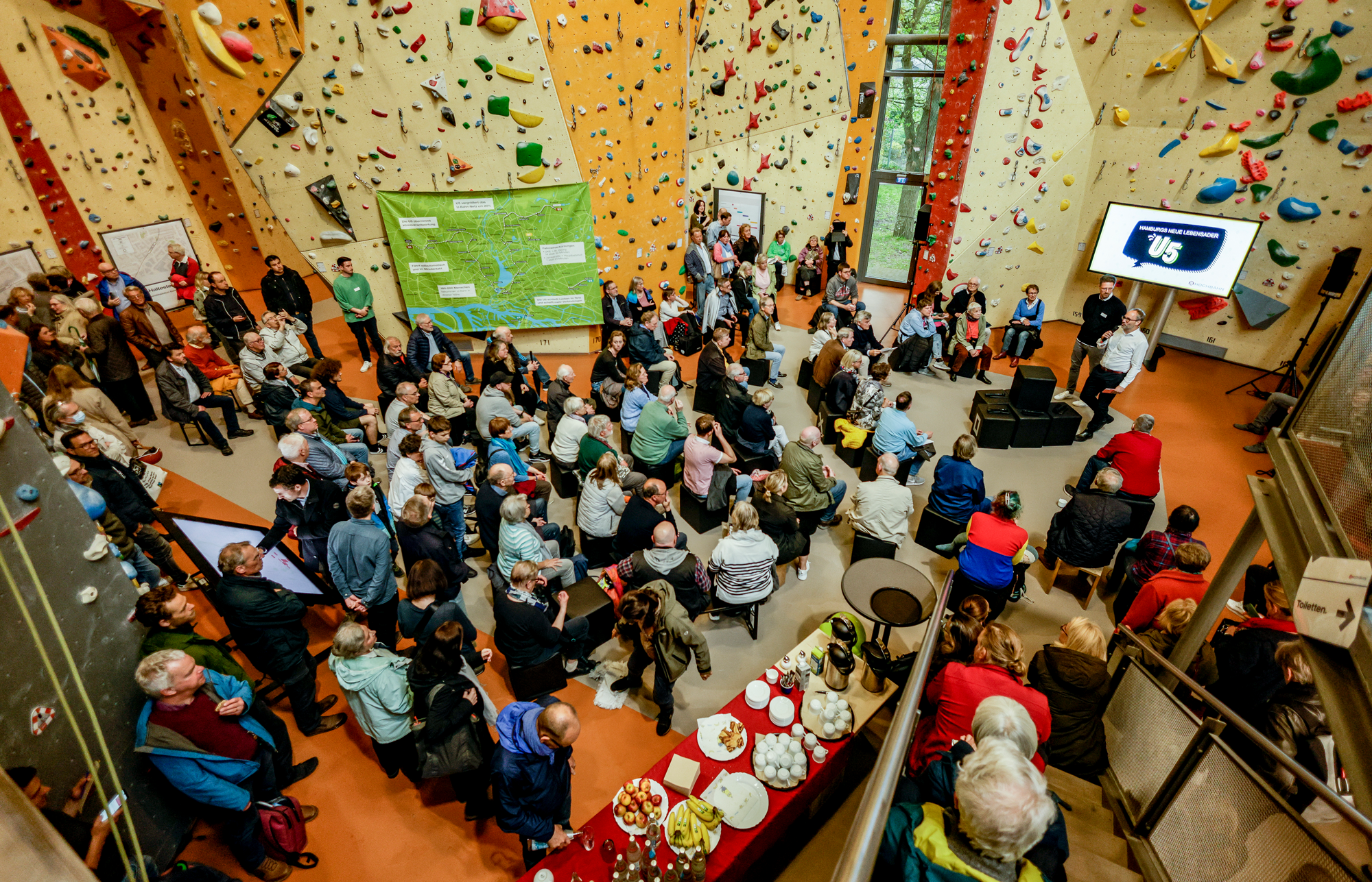 Foto einer Veranstaltung in einer Kletterhalle von oben, zeigt Publikum, Infografiken, Präsentation