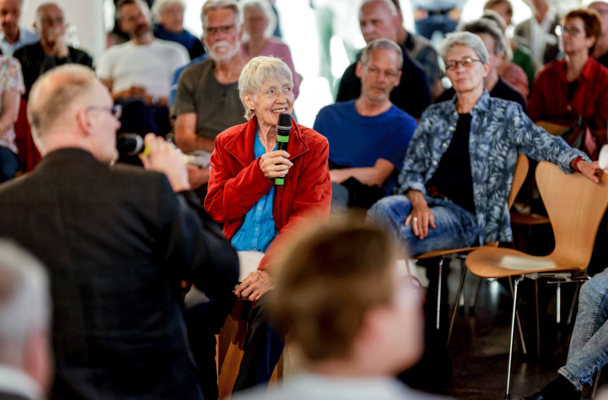 Bild bei einer Veranstaltung, im Fokus eine Frau mit Mikro, im Hintergrund weiteres Publikum.