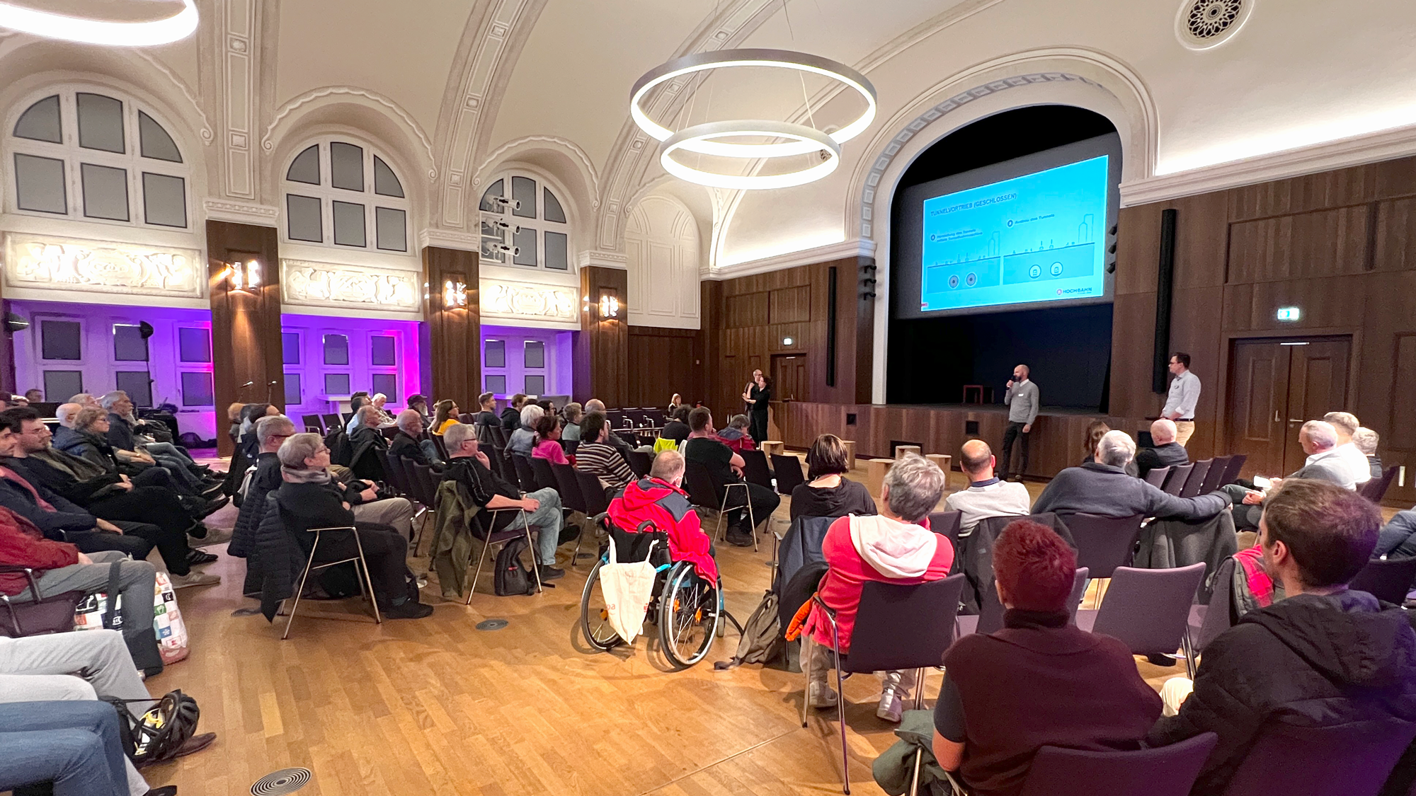 Veranstaltungsaal mit Publikum, welches einer Präsentation zuhört.