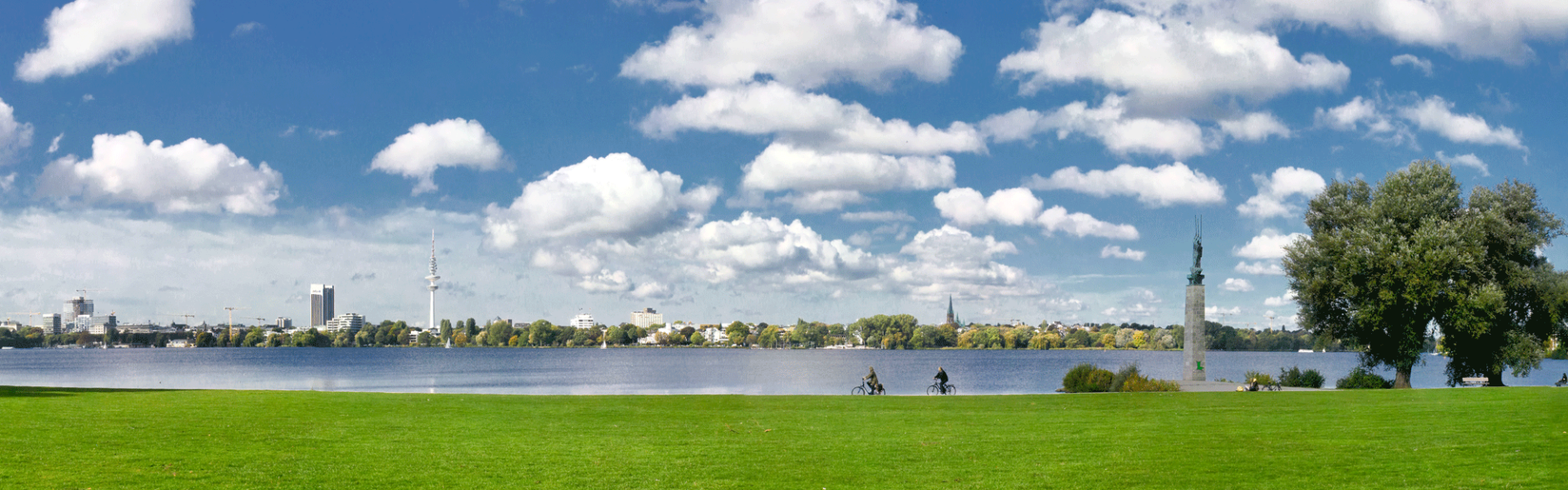 Panoramabild der Alster. Im unteren Teils des Bildes, strahlend grünes Gras, darüber die Alster ind ein blauer Himmel mit kleinen Wolken. Am Horizont Teile der Hamburg Skyline. 