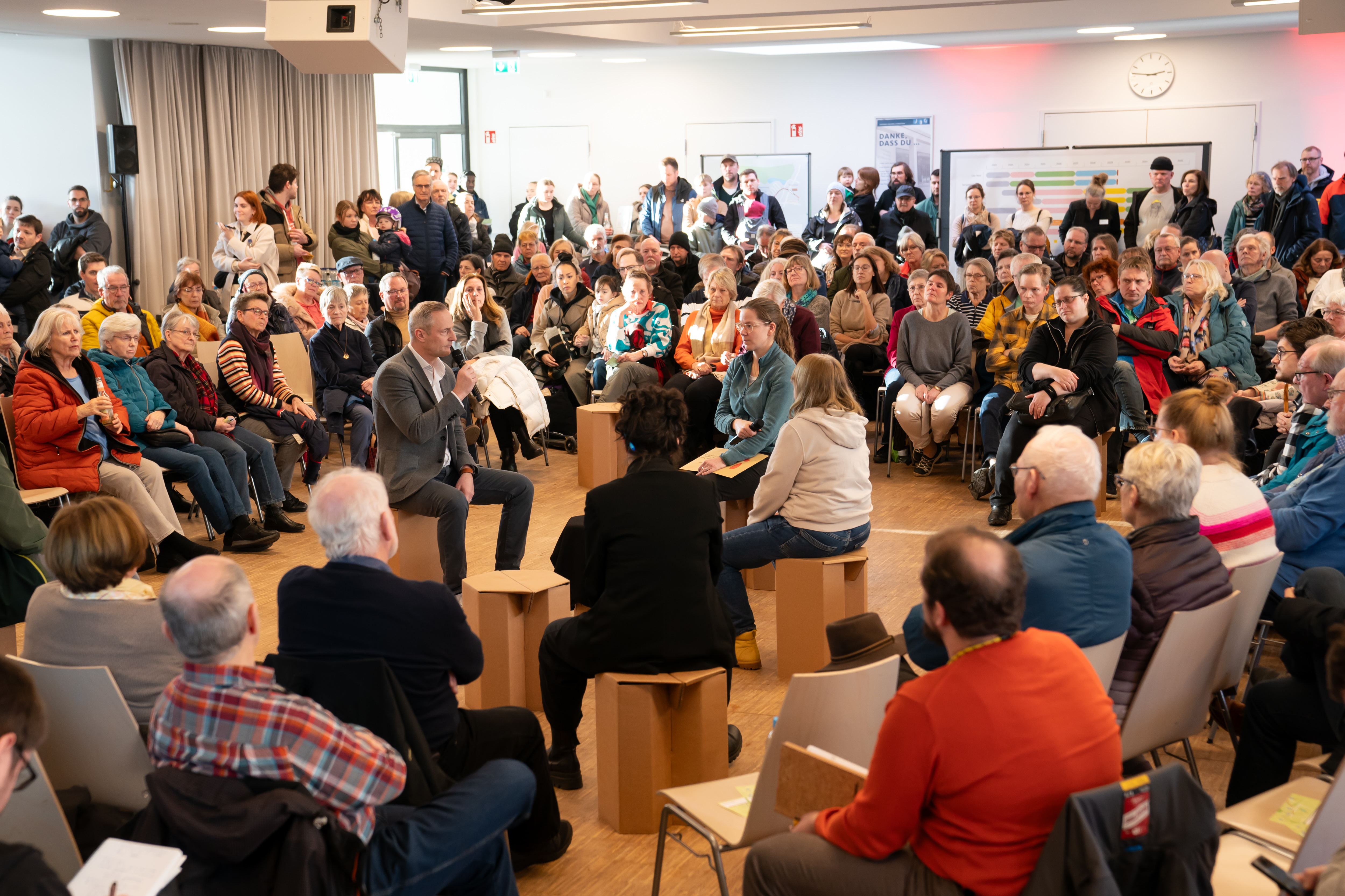 Menschen sitzen im Kreis um mehrere Hocker in der Mitte, in der Personen mit Mikrophon sitzen. 