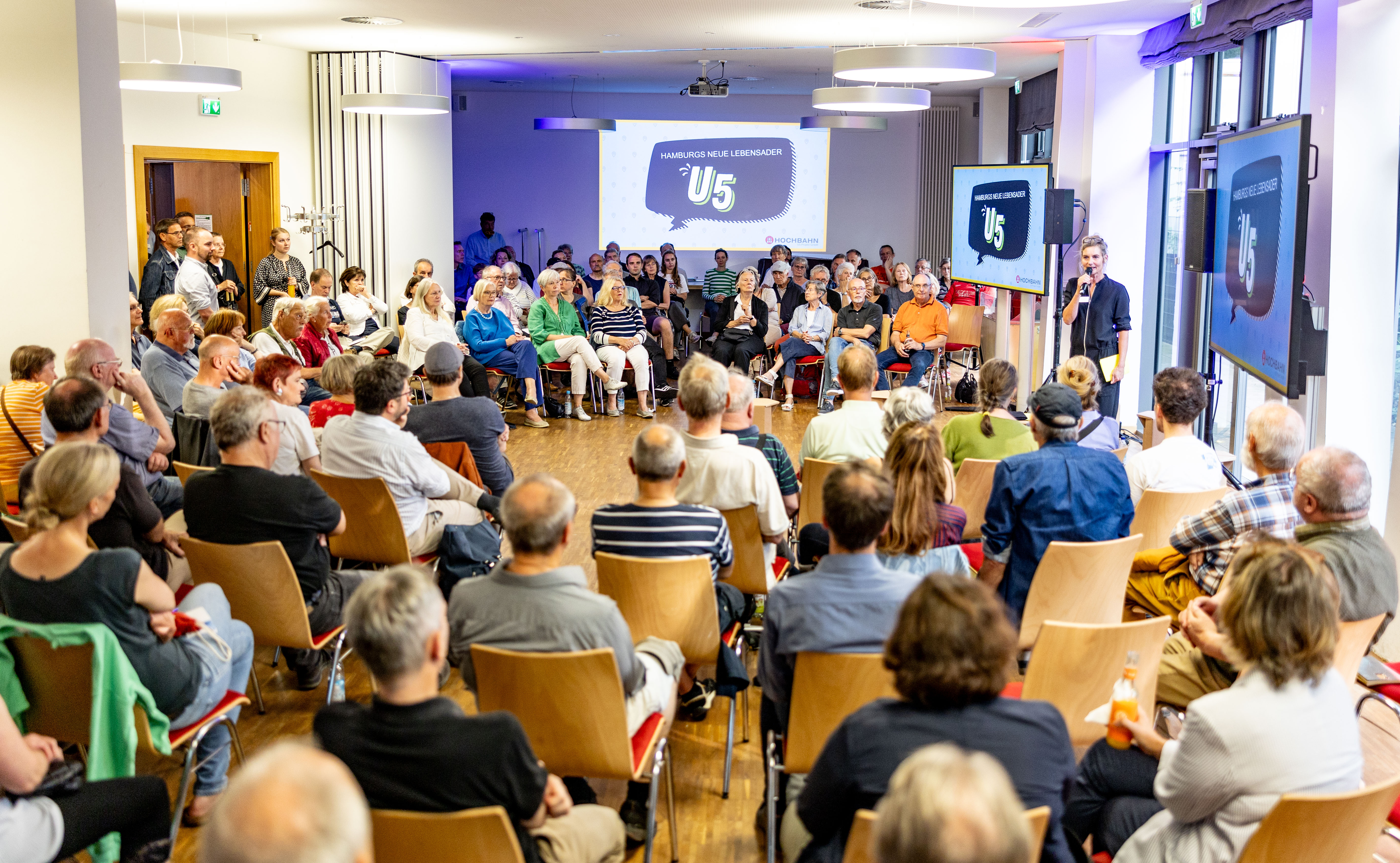 Publikum sitzt im Kreis um einen Redner, links und rechts zwei Präsentationen. 