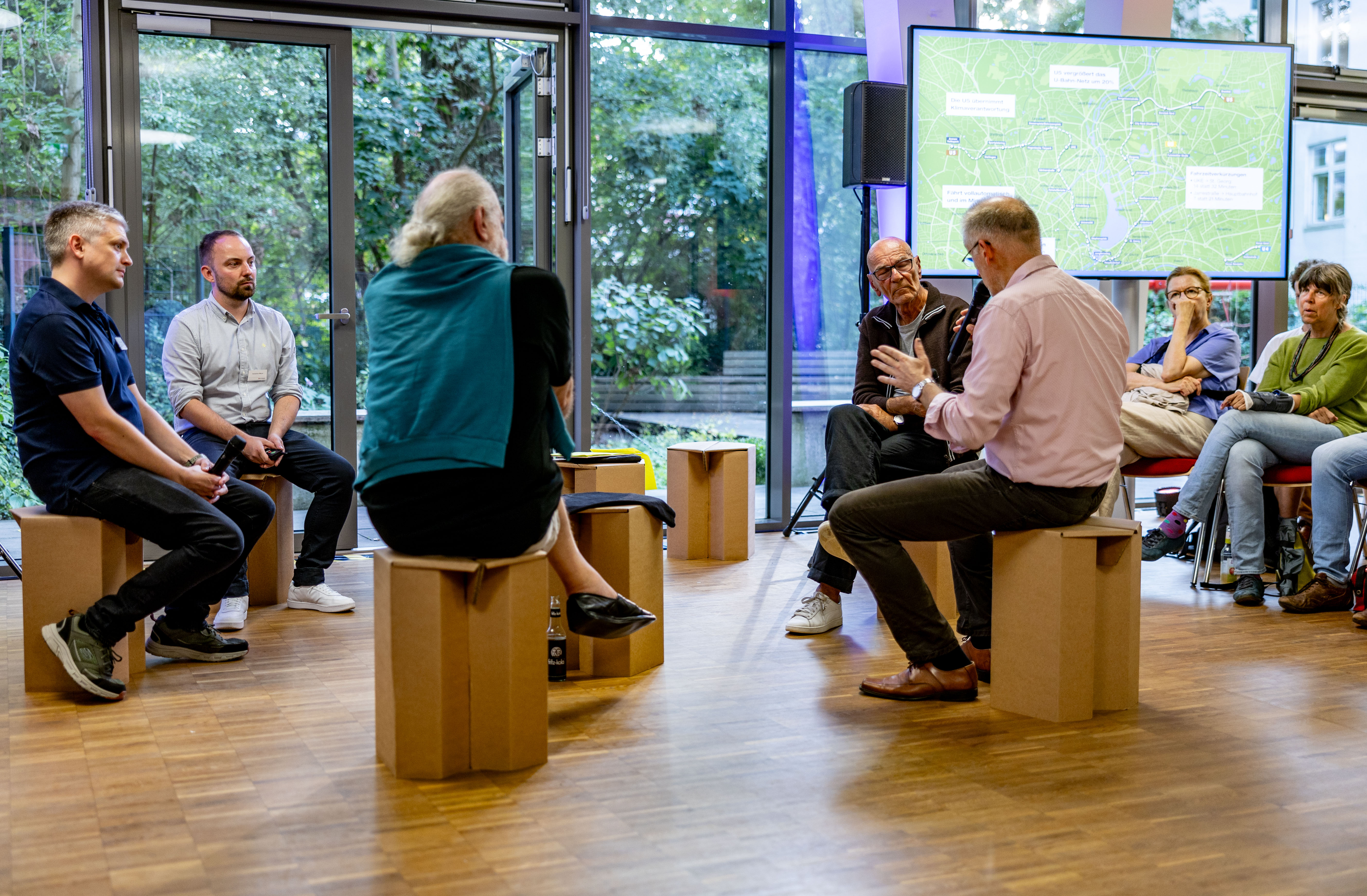 Die Fachleute des U5-Projektteams sitzen bei einer Veranstaltung mit Interessierten im Kreis. Im Hintergrund eine Präsentation und weitere Zuhörende.