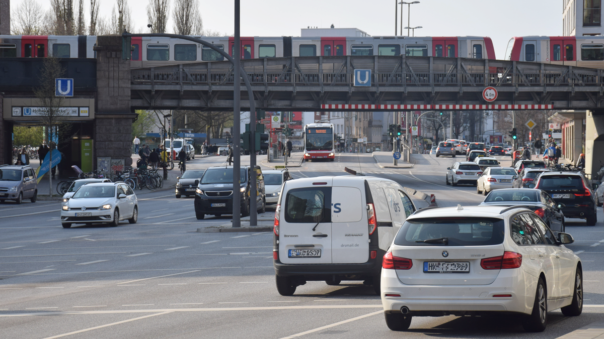 Mehrspurige befahrene Straße. Im Hintergrund die Haltestelle U-Hoheluftbrücke. Auf der Brücke eine Bahn.