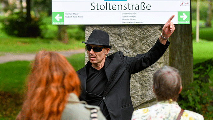 Ein Schauspieler mit Hut, Sonnenbrille und schwarzem Anzug steht beim Spaziergang „Horn-to-go“  vor einem Haltestellenschild mit der Aufschrift "Stoltenstraße".