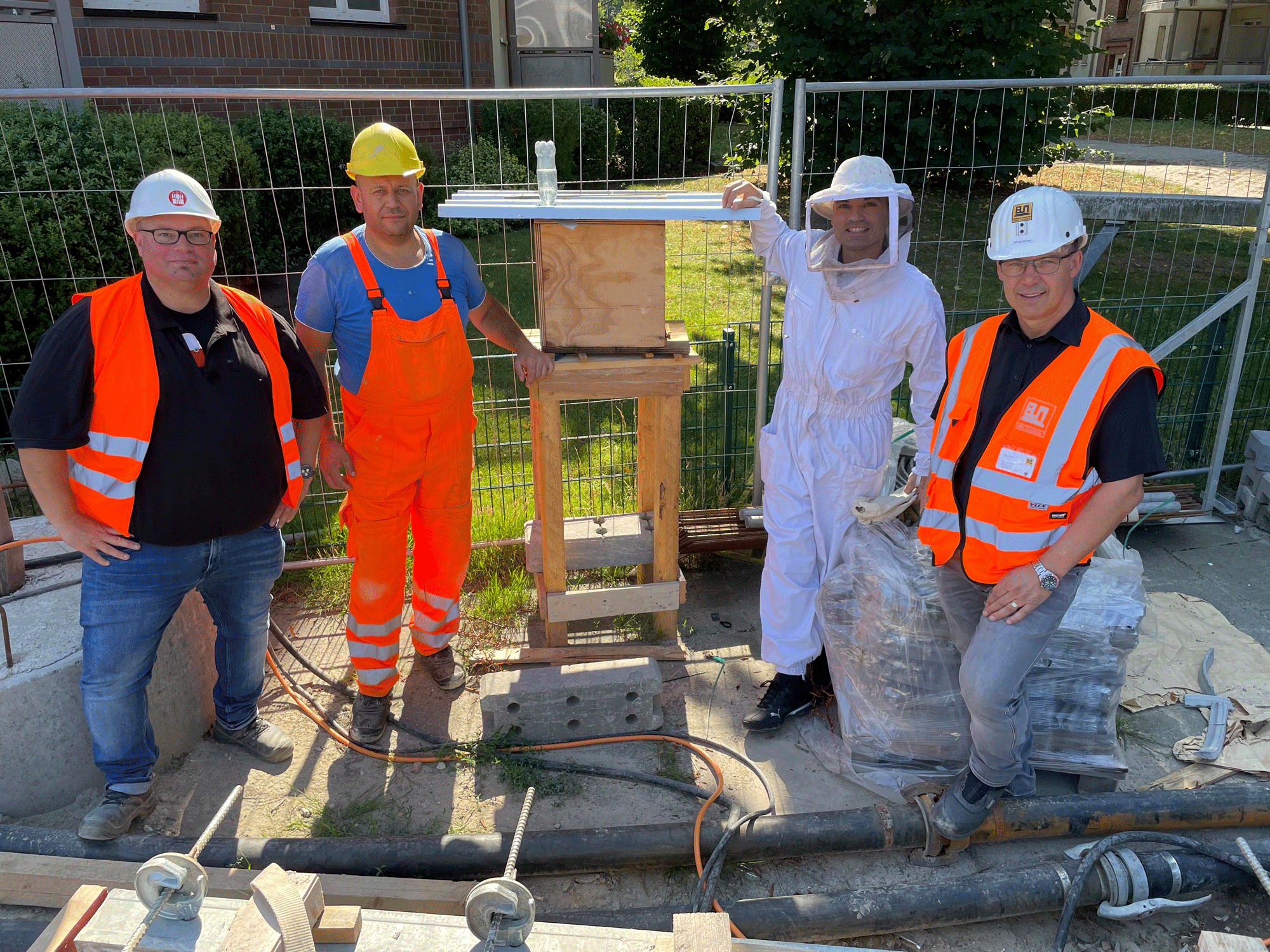 Vier Personen, davon 3 in Sicherheitskleidung für die Baustelle und eine in Imker-Kleidung, posieren neben einem Holzkasten 