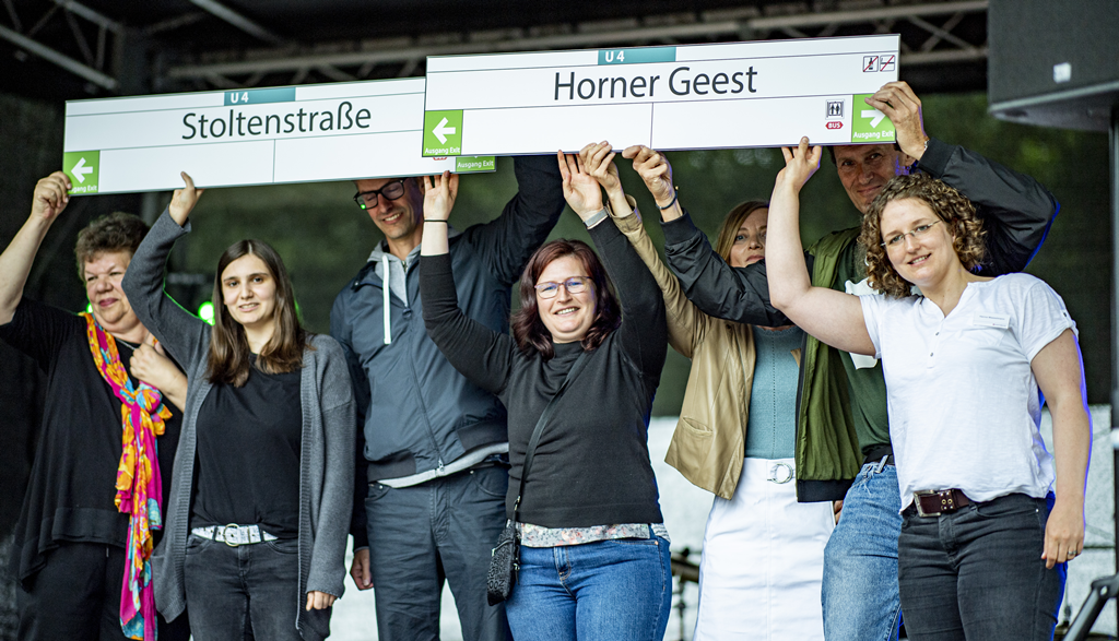 Die Mitglieder der Jury halten Haltestellenschilder mit der Beschriftung "Horner Geest" und "Stoltenstraße" über ihre Köpfe.