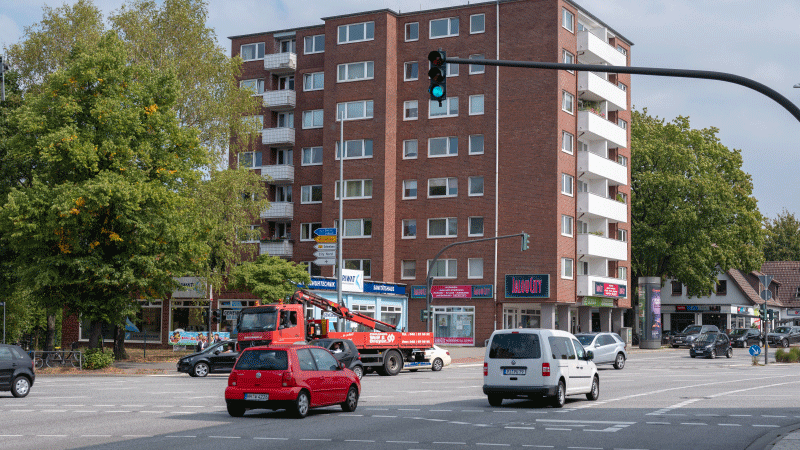 Mehrspurige befahrene Straße. Im Hintergrund ein mehrstöckiges Wohnhaus. 