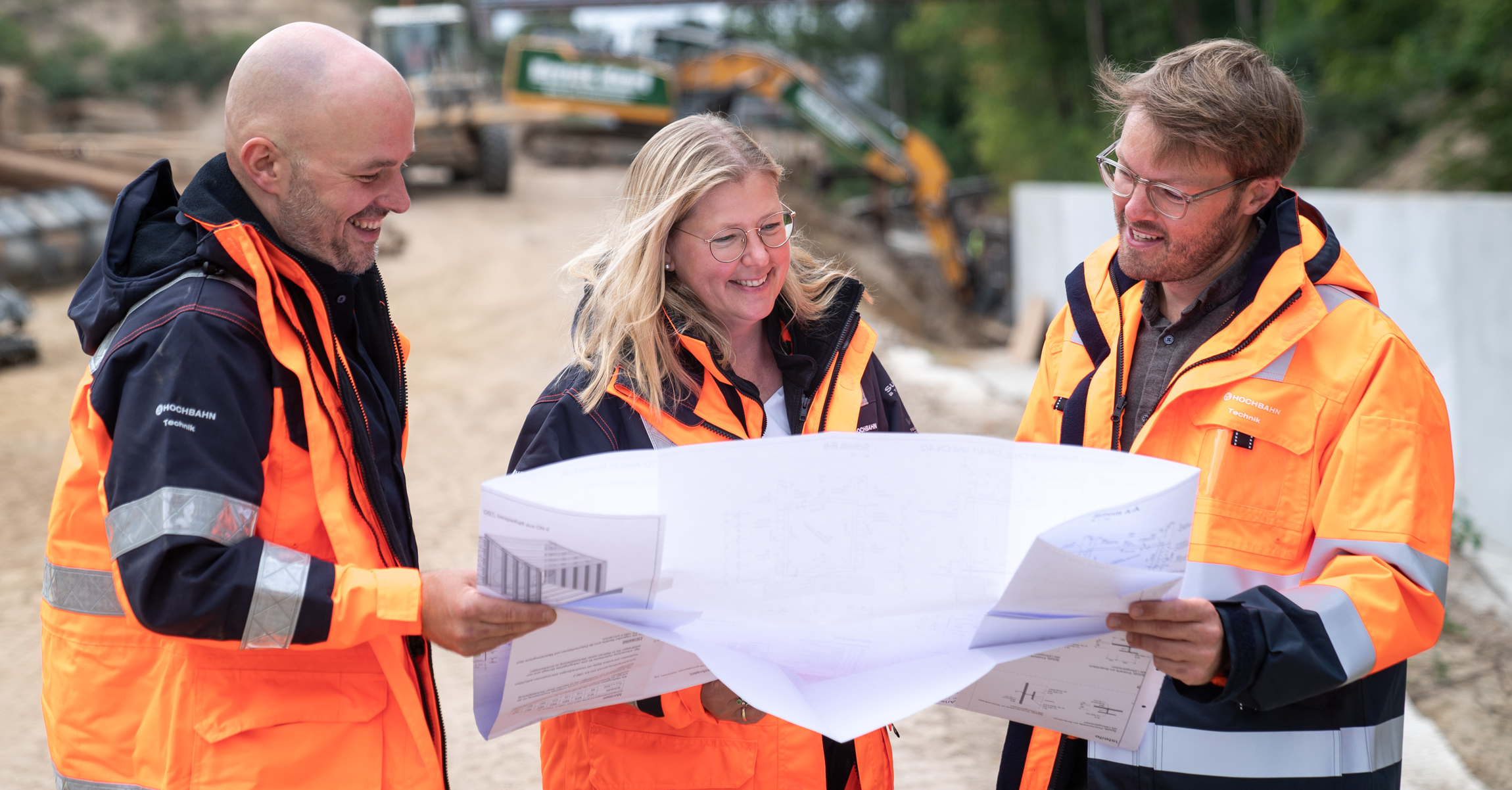 Drei HOCHBAHN-Mitarbeiter*innen stehen auf der Baustelle und schauen lächeln gemeinsam auf einen Plan