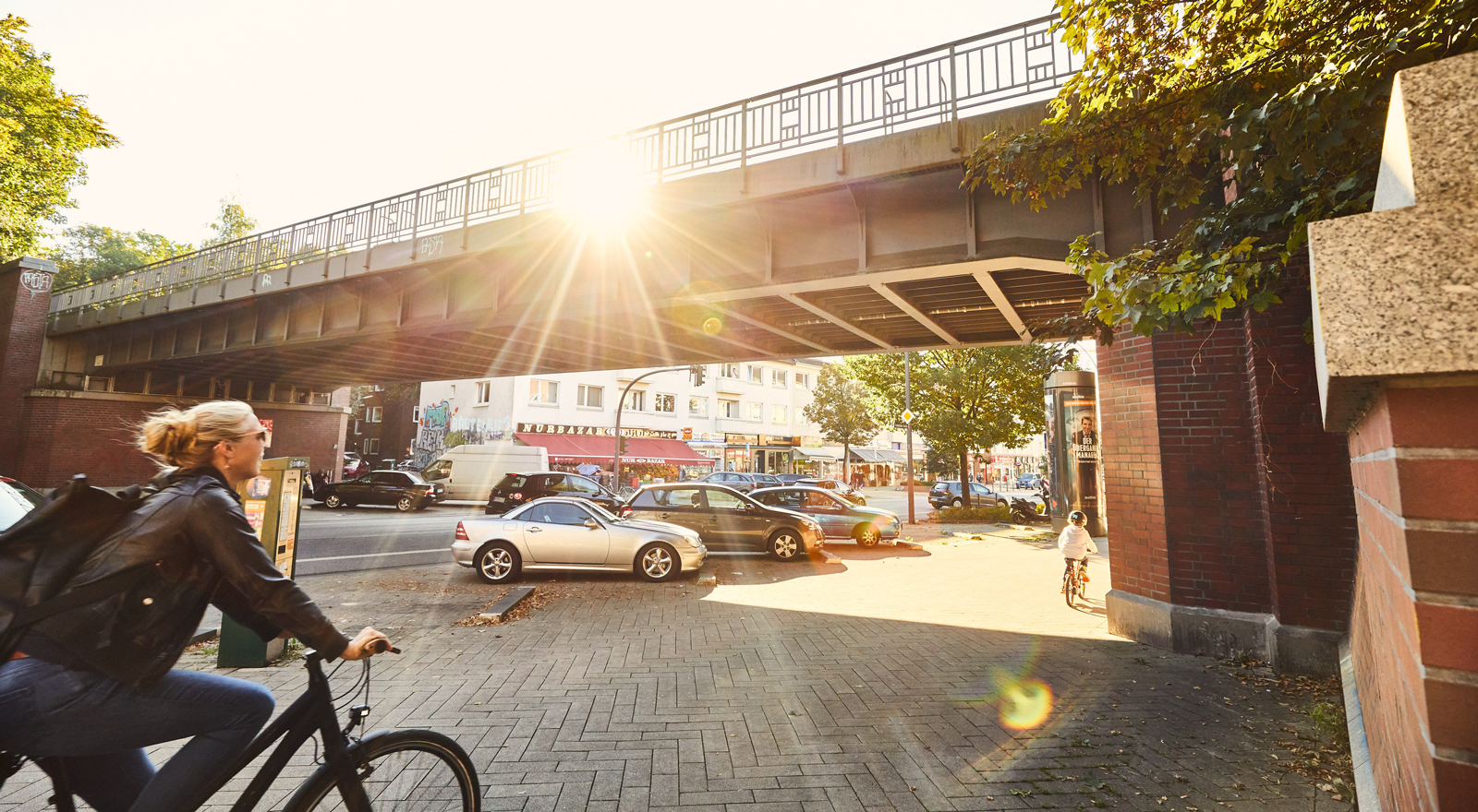 Bild vom Bürgersteig auf die Brücke der U3 über die Fuhlsbüttler Straße. Autos parken unterhalb der Brücke, die Sonne scheint, eine Frau aufm Fahrrad fährt durch das Bild. 