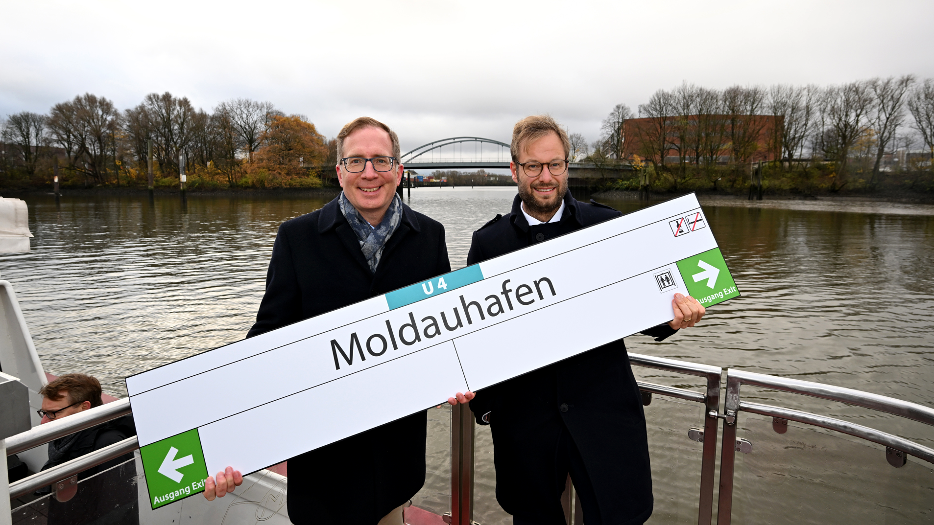 Zwei Personen halten Haltestellenschild mit der Aufschrift "U4 Moldauhafen" im Arm. 