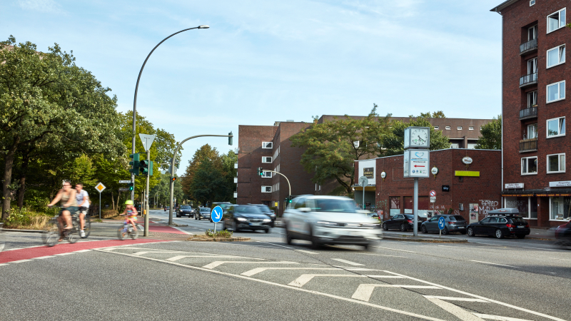 Straße mit Autos und Fahrradfahrern. Auf einer Straßenseite, mehrstöckige Wohnhäuser