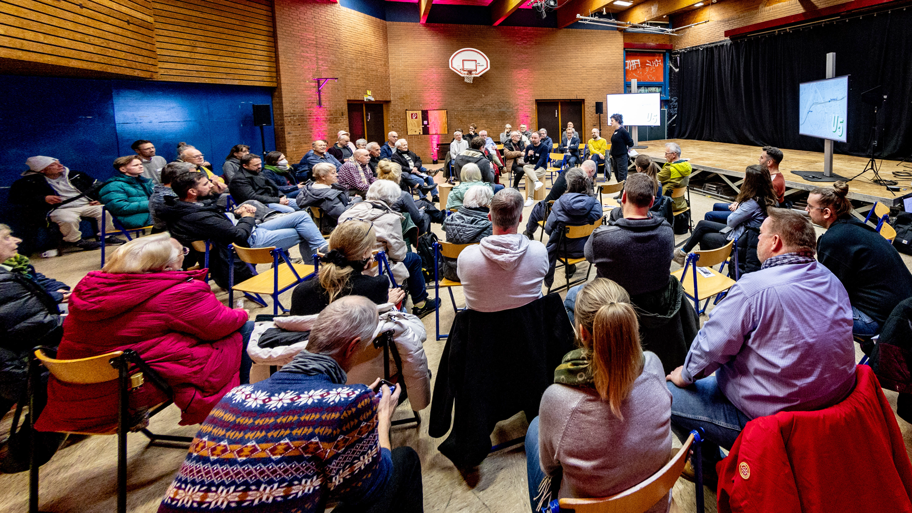 Veranstaltungsraum in dem einige Menschen dem Redner in der Mitte zuhören. Links und Rechts jeweils ein Bildschirm mit einer Präsentation.