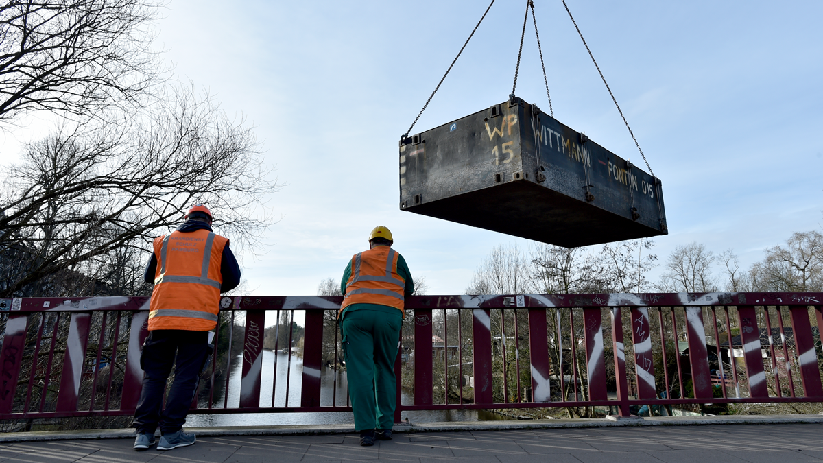 Zwei Personen mit Warnwesten und Helm stehen an einer Brücke. Eine Insel wird von einem Kran ins Wasser gelassen