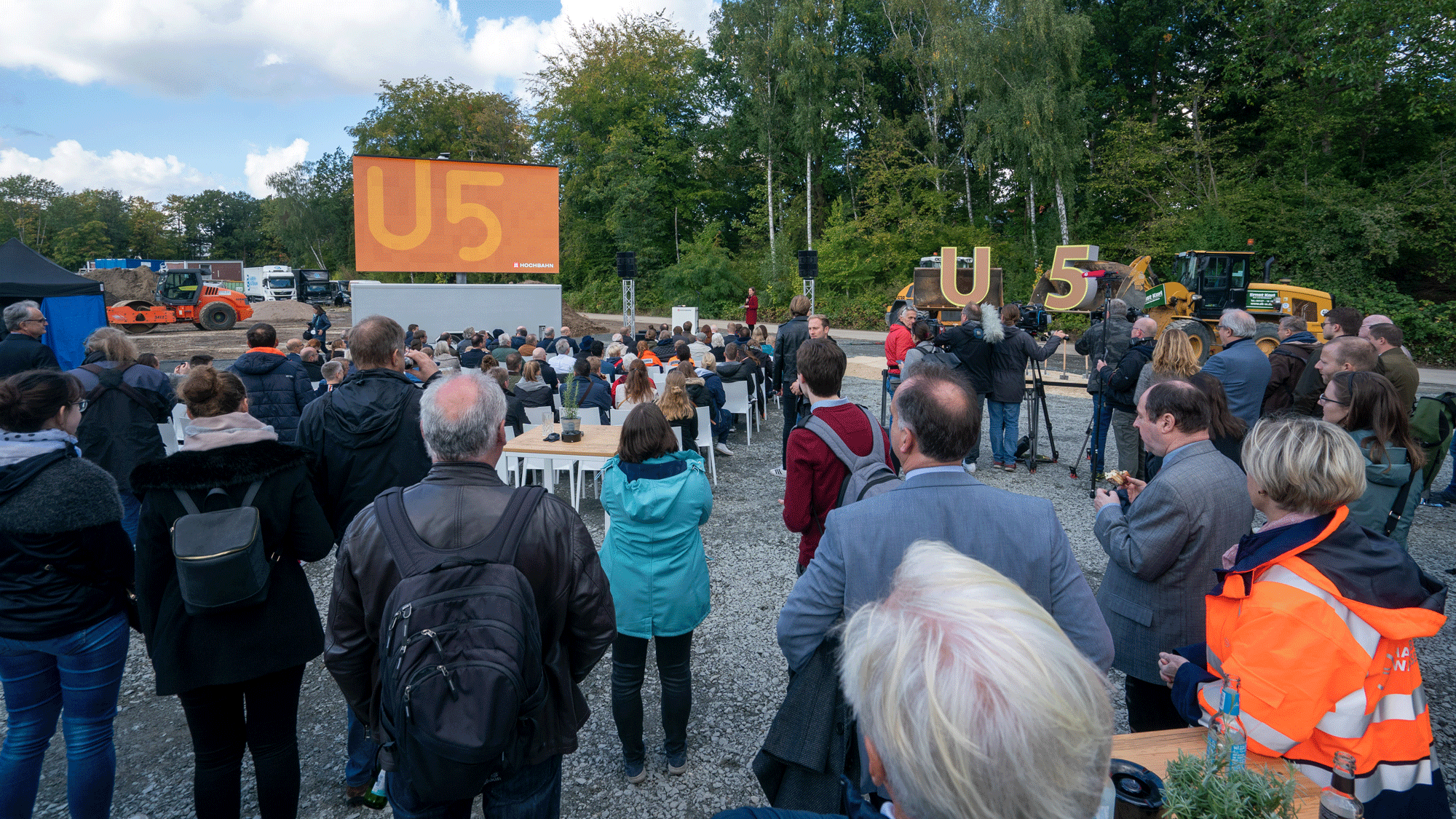 Eine große Gruppe von Menschen steht auf der Baustelle für den ersten Spatenstich und schaut auf eine Leinwand.