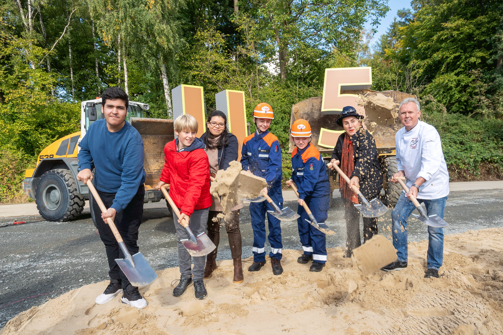 Eine Gruppe von Menschen mit Spaten vor zwei Baggern die in ihren Schaufeln ein großes U und eine große 5 haben.