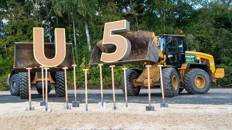 Zwei Bagger haben ein großes U und eine große 5 in ihrer Baggerschaufel. Im Vordergrund sind mehrere Spaten in der Erde.