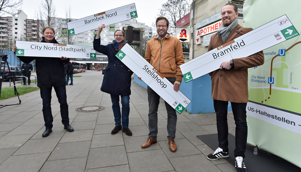 Vier Personen halten Haltestellenschilder mit der Beschriftung "Bramfeld", "Steilshoop",  "Barmbek Nord", und "City Nord (Stadtpark)" in ihren Armen.