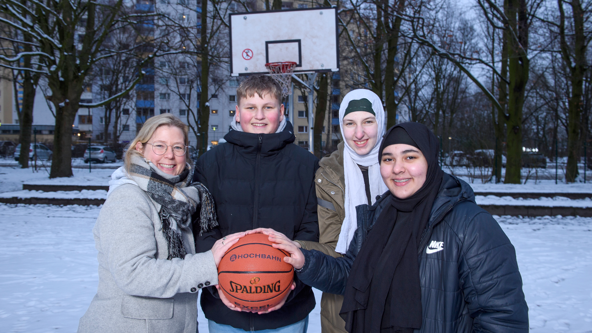 U5-Beauftragte Tanja Taaks mit vier Jugendlichen aus Steilshoop auf einem Basketballplatz. Alle haben ihre Hände auf einen Basketball gelegt. 