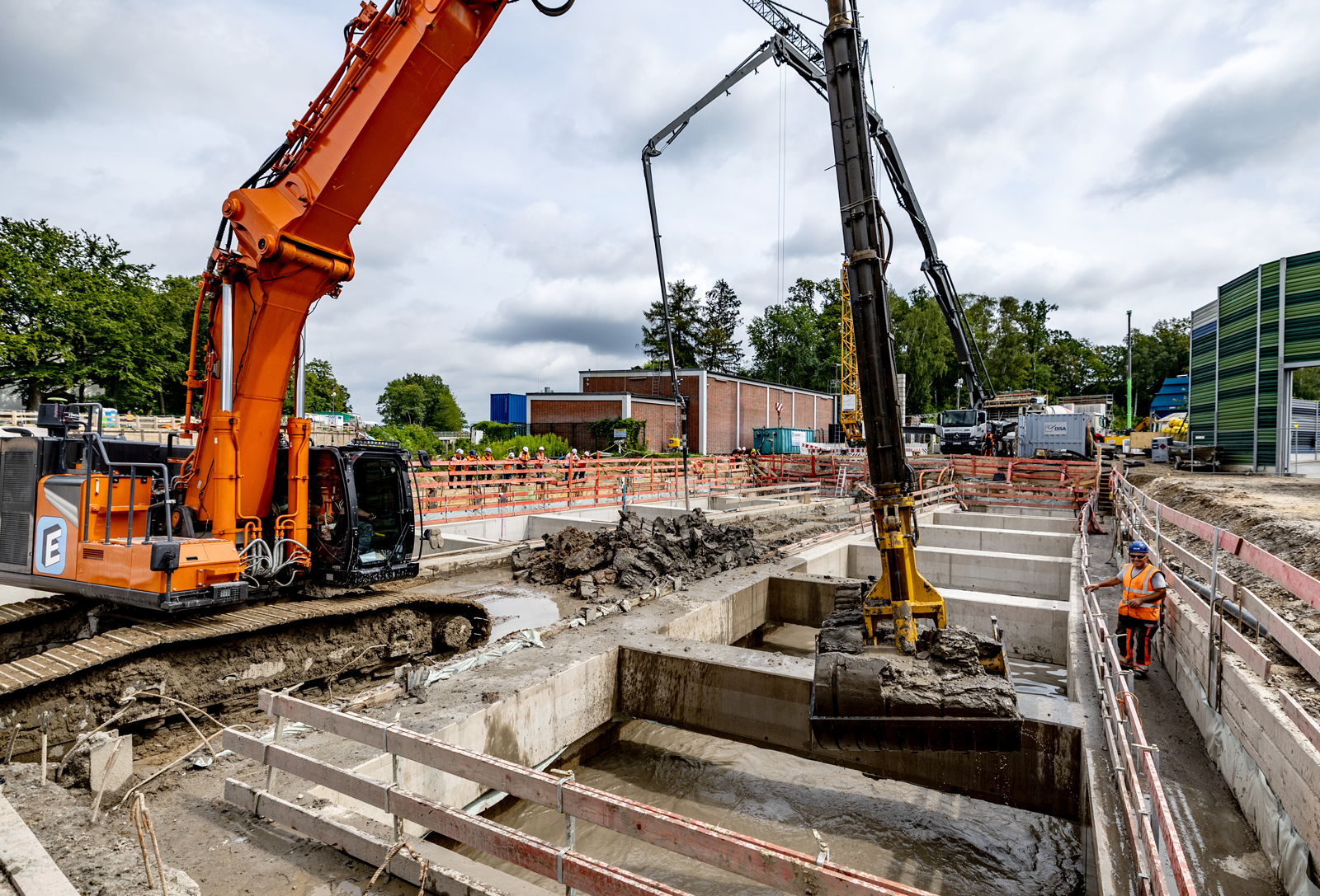 Bagger hebt Erde aus einer Baugrube mit Wasser aus, im Hintergrund der Busbetriebshof Alsterdorf 