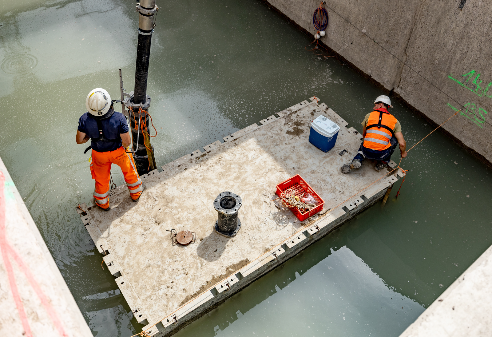 Blick von oben auf ein Ponton in der Baugrube. Zwei Personen stehen darauf mit einem großen Schlauch in der Hand. 