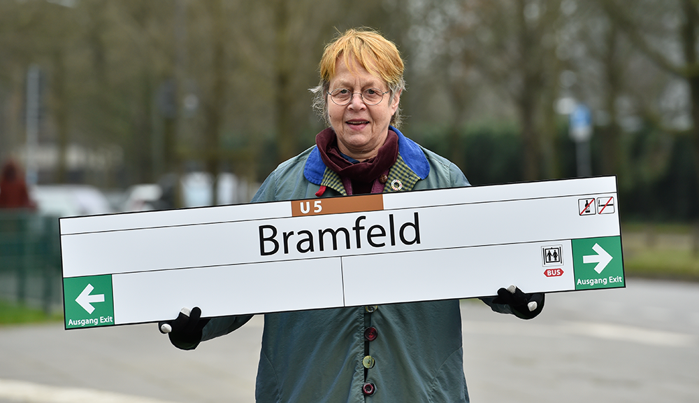 Eine Frau hält ein Haltestellenschild im Arm mit der Aufschrift Bramfeld