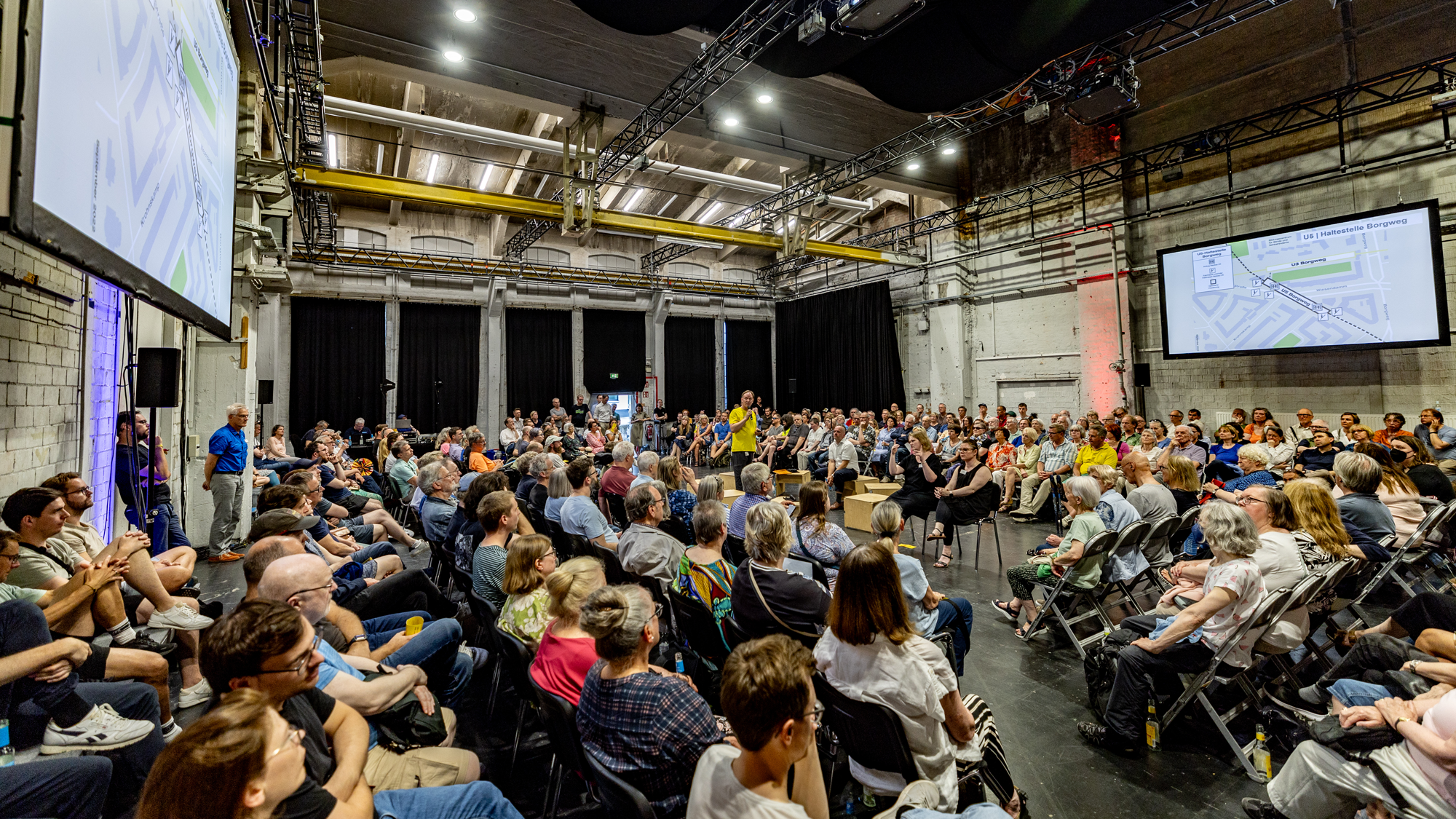 Eine Veranstaltungshalle ist gefüllt mit Menschen. Sie sitzen in Stuhlreihen im Kreis um eine Diskussionsrunde in der Mitte des Raumes. Links und rechts an den Wänden, große Leinwände mit einer Präsentation. 