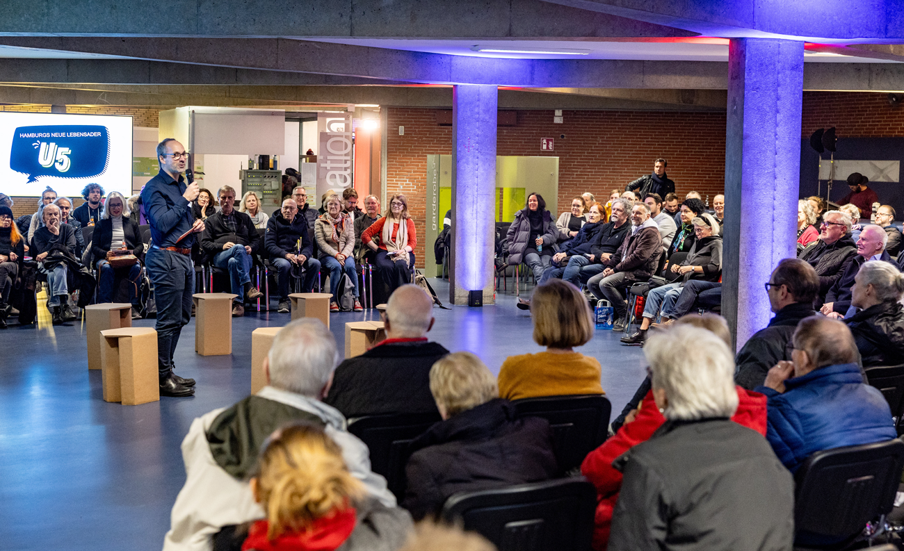 Raum mit Menschen, die in einem Halbkreis um den in der Mitte stehenden Sprecher sitzen. Neben dem Sprecher leere Hocker 