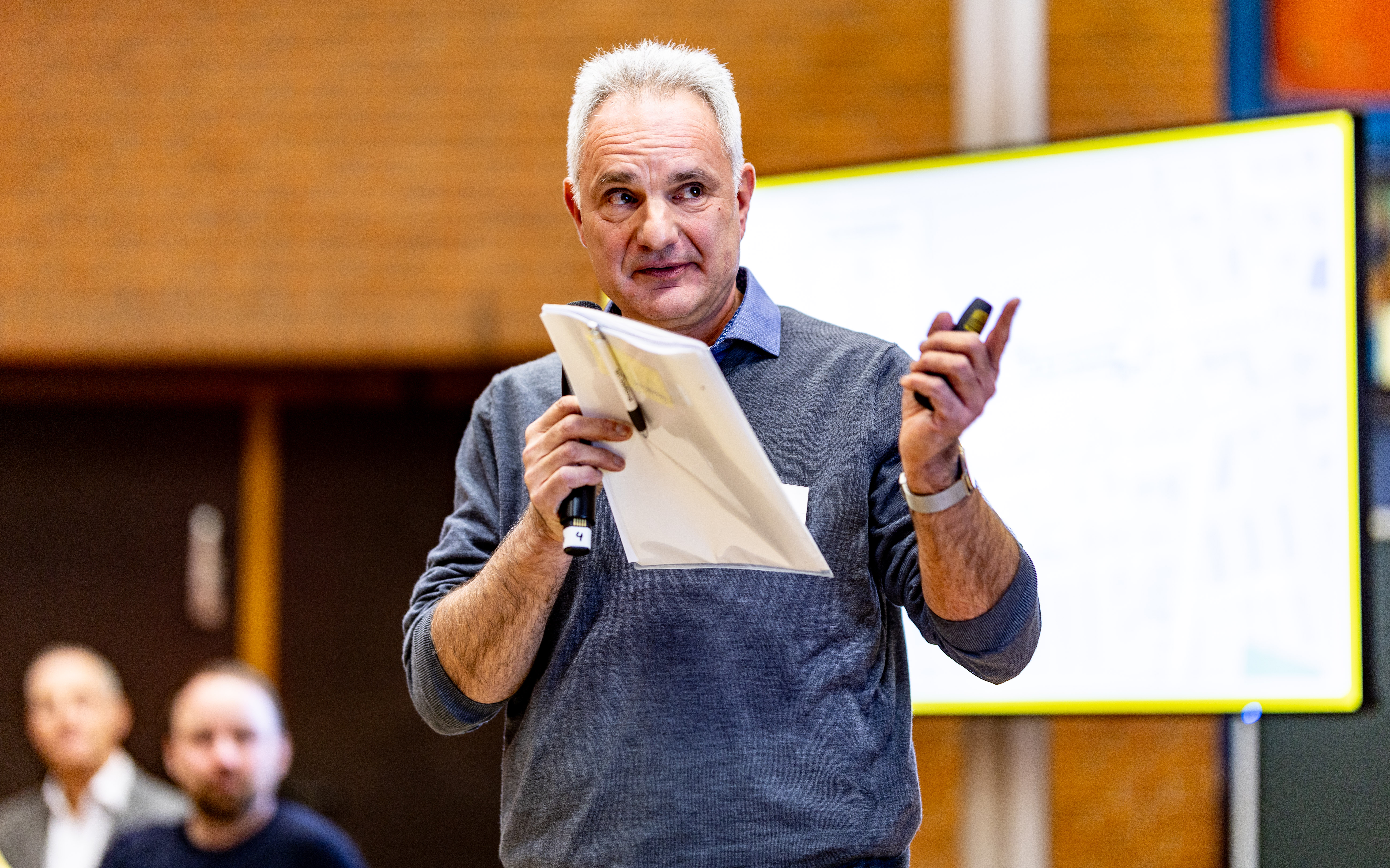 Ein Mann mit Mikro und einer Mappe in der Hand steht vor einer Präsentation. 