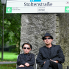 Ein Kind und ein Mann, beide in schwarzen Anzügen und mit Sonnenbrille, sitzen untern einem Haltestellenschild mit der Aufschrift "Stoltenstraße".